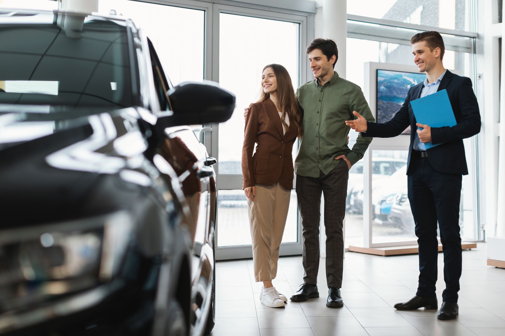 Happy car salesman showing used automobile to young couple at auto dealership